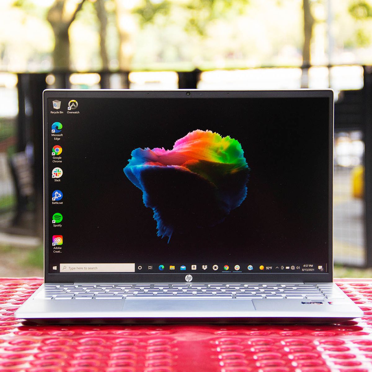 The HP Pavilion Aero 13 open on a red picnic table with a playground in the background. The screen displays a black desktop background with a multicolored cloud in the center.