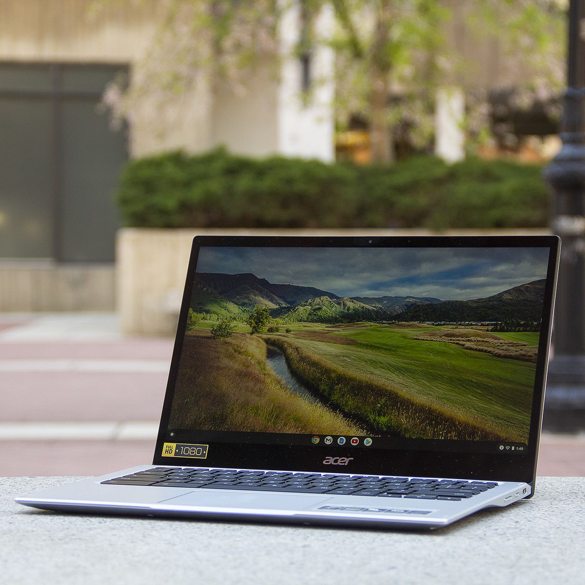 The Acer Chromebook Spin 513 sits on a stone bench, open, angled to the left. The screen displays a pastoral scene.