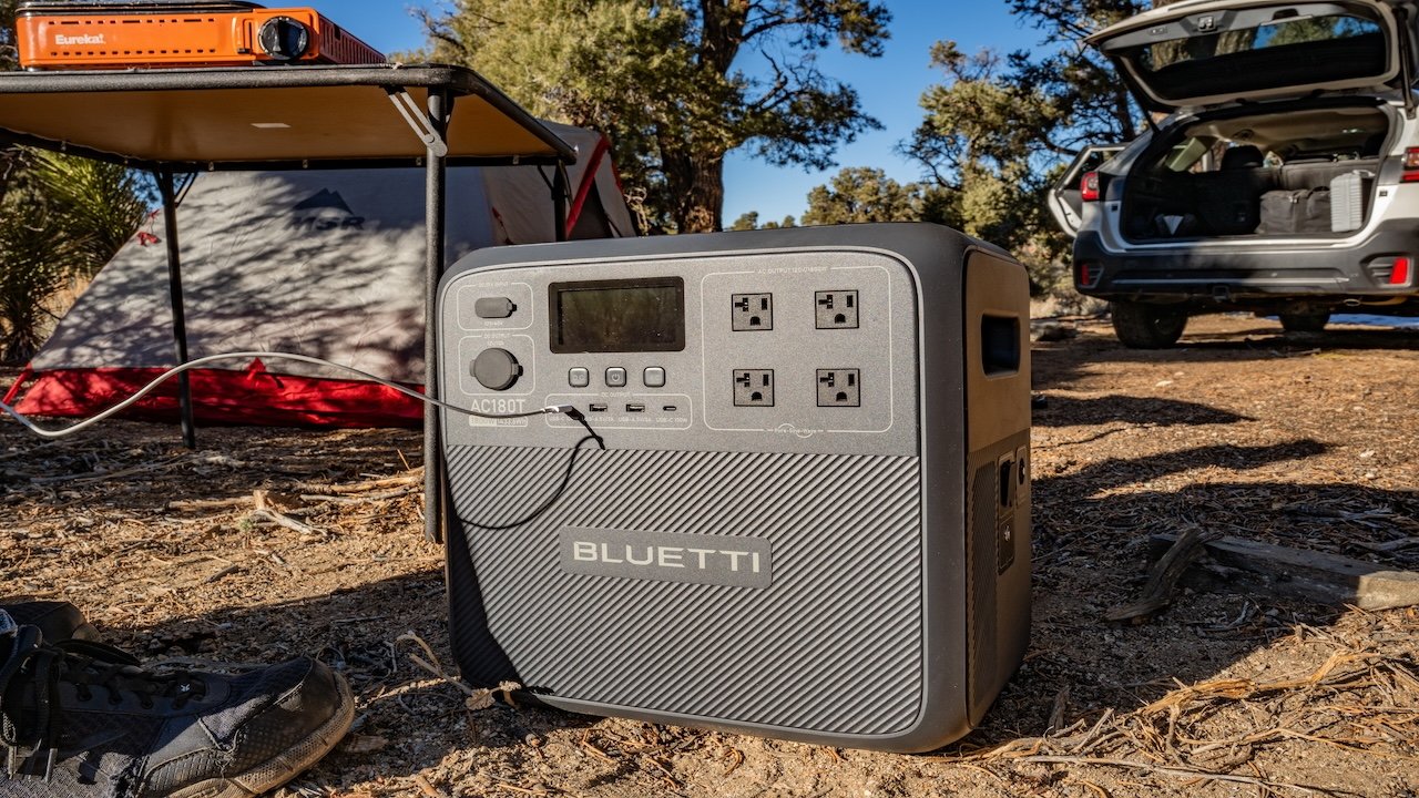 A portable power station on the ground with outdoor camping gear and an open car trunk in the background.