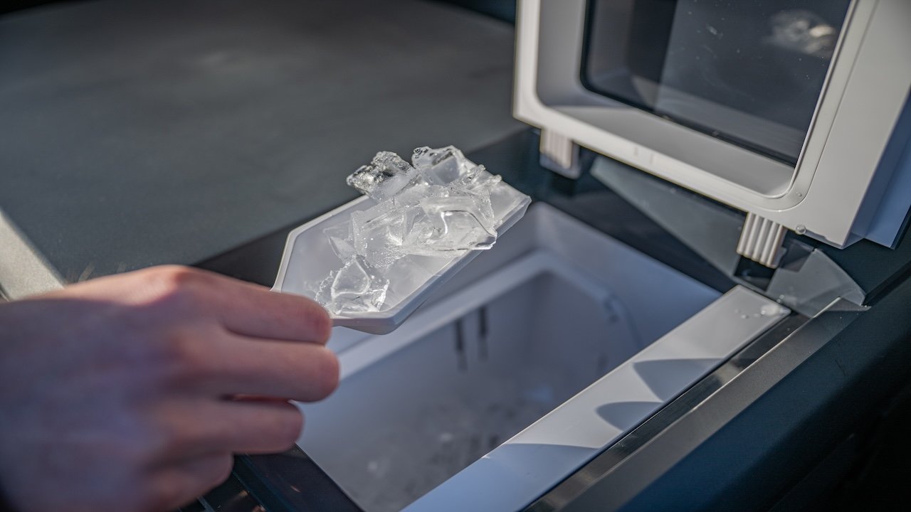 A hand holding a tray of ice cubes over an open freezer compartment, ready to deposit them.