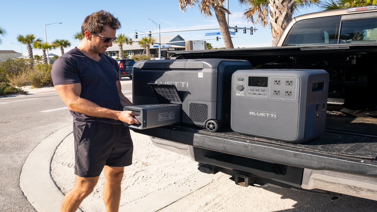 Man unpacking a portable power station from the back of a pickup truck on a sunny day with palm trees in the background.