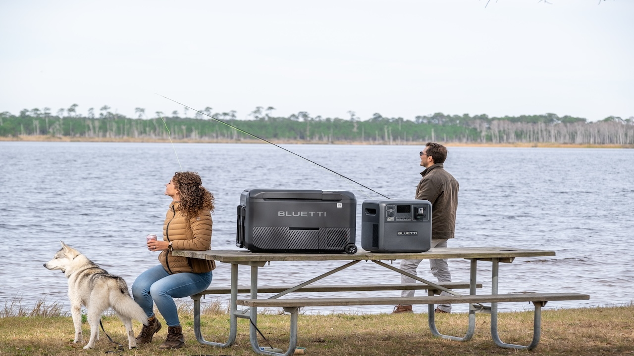 Man and woman with a dog sitting at a picnic table by a lakeside, with portable power stations on the table.