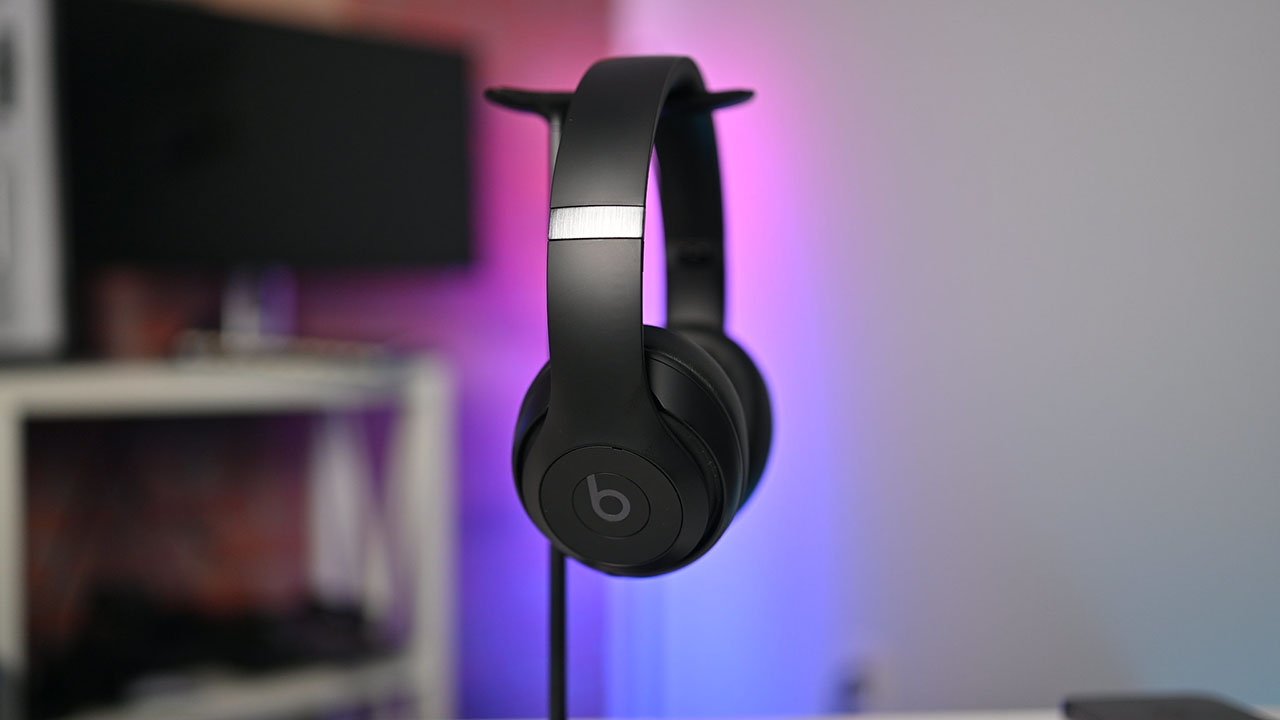 Beats Studio Pro headphones hanging on a stand, with blurry monitors and purple lighting in the background.