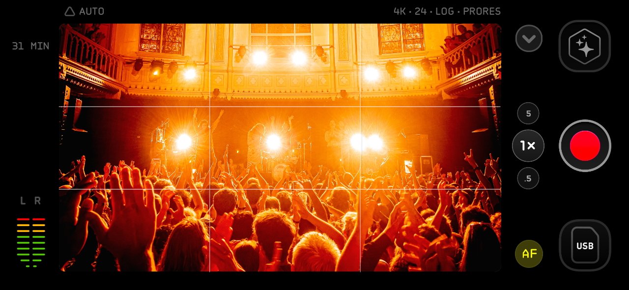 Large crowd at a concert with bright stage lights and performers in a grand hall, viewed through a camera screen interface.