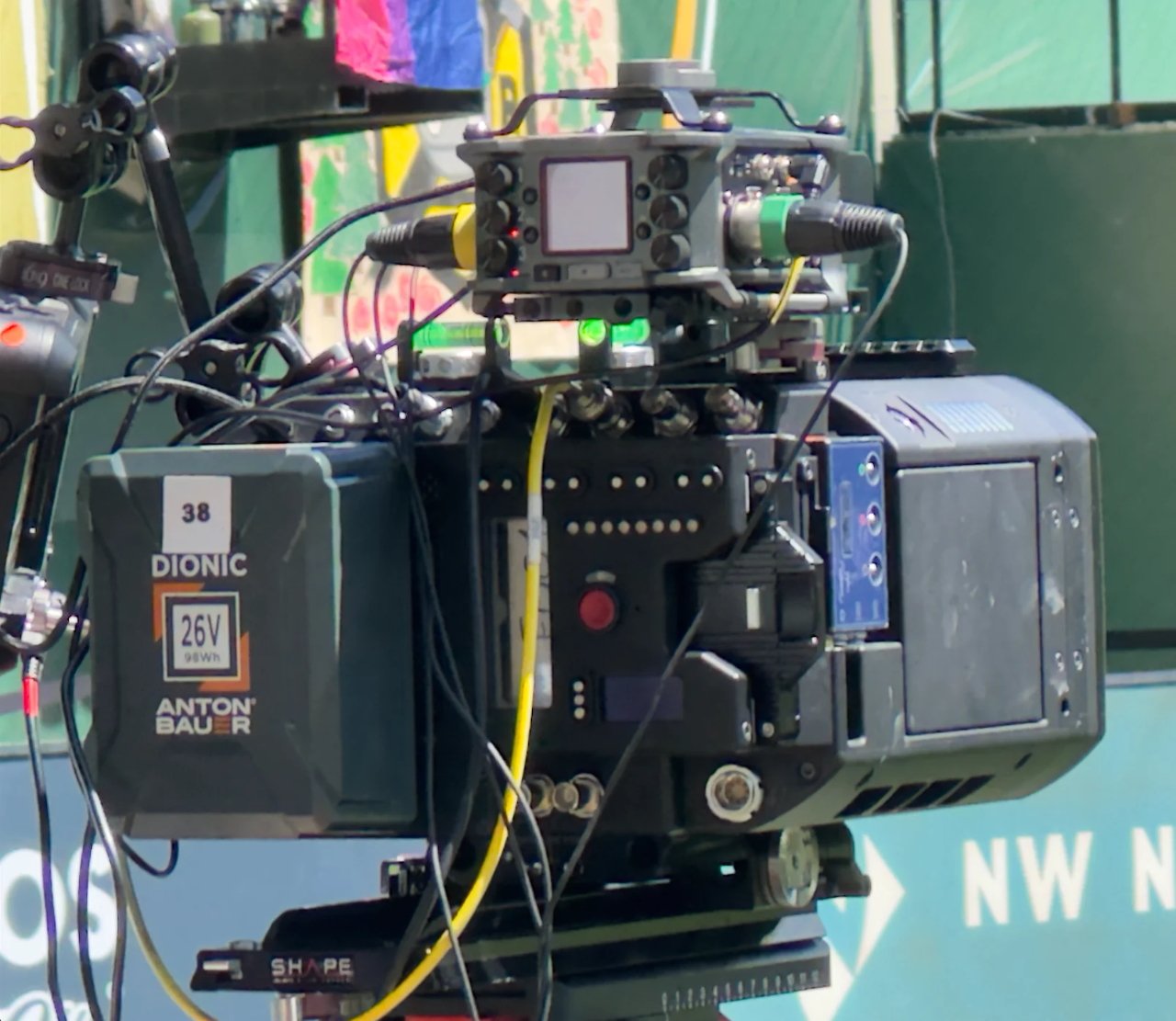 Close-up of a professional video camera with multiple cables, battery pack, and control panel. Background includes colorful posters and a green wall with the letters 'NW' visible.