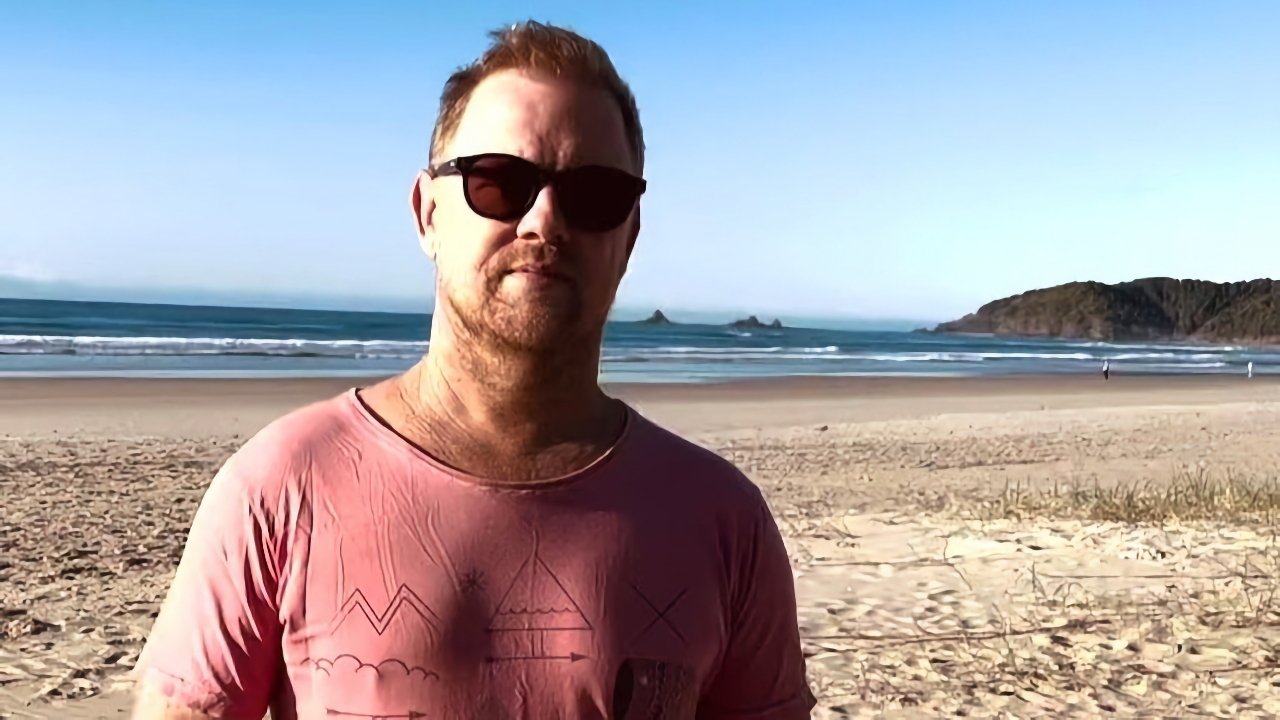 Man in sunglasses and pink shirt stands on a sandy beach with ocean and distant islands in the background.