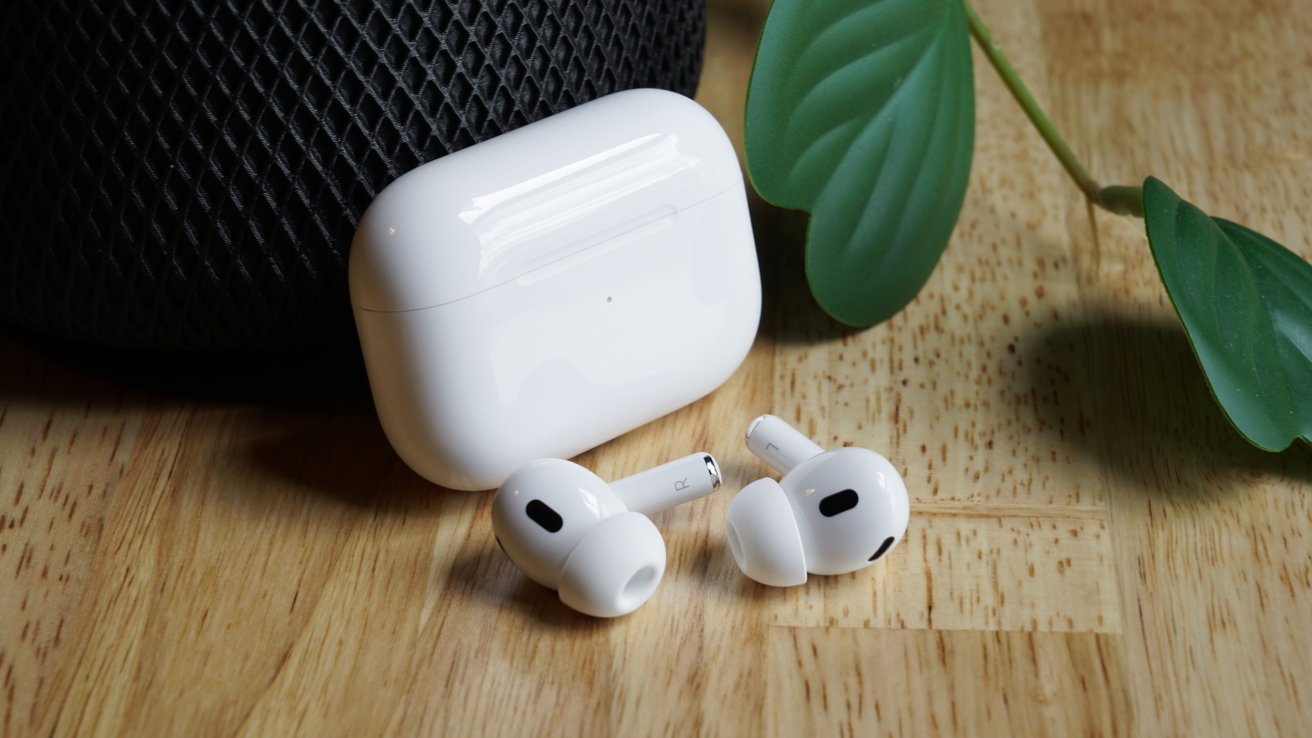 AirPods next to their open charging case on a wooden surface, with a plant leaf and HomePod speaker in the background.