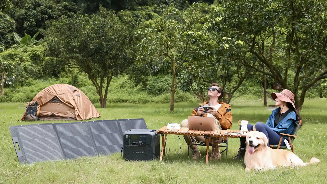 Two people and a dog relax at a campsite with a tent, solar panels, and a table with a laptop and camping gear.