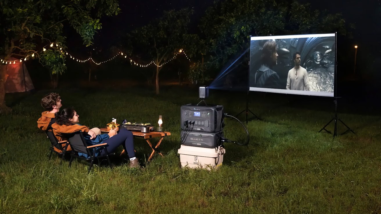 Two people sitting on chairs outdoors watching a movie projected on a screen, surrounded by trees and string lights at night.