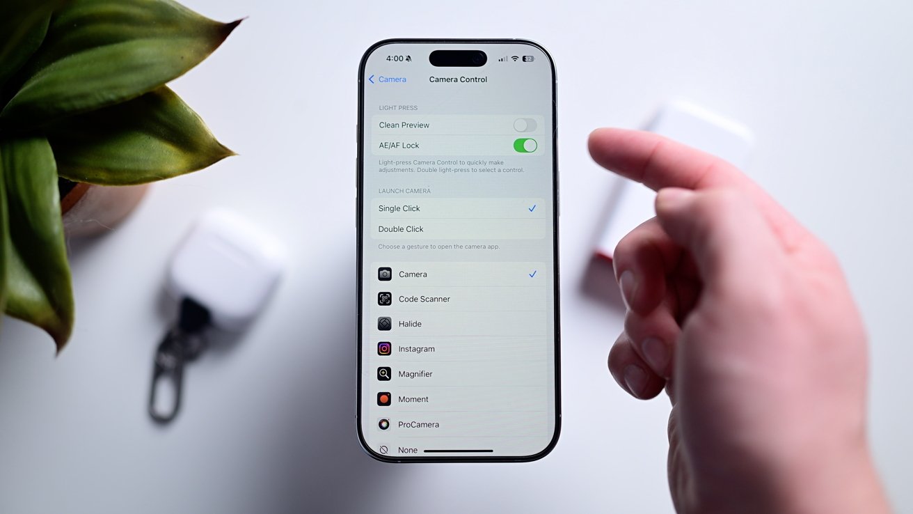 Hand holding a smartphone displaying camera control settings, near a plant and wireless earbuds case on a white surface.