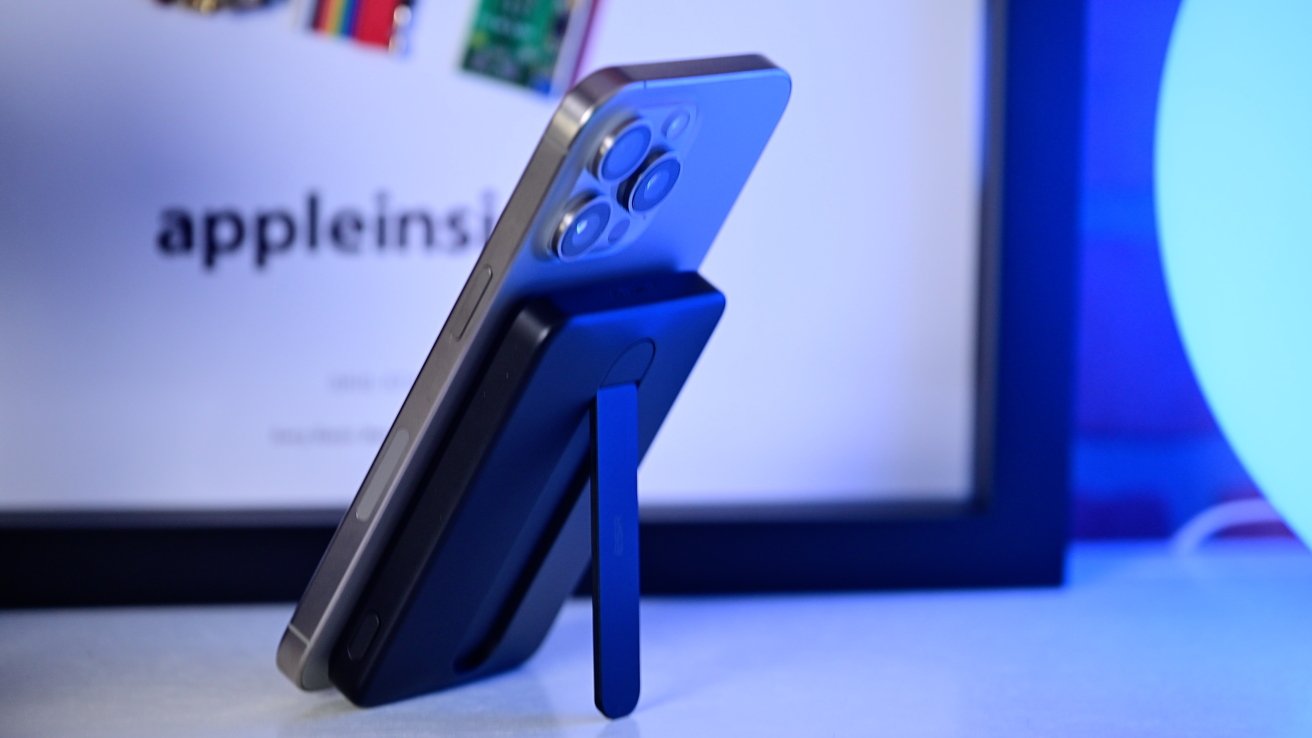 Smartphone with three cameras stands upright on a table, supported by a blue portable battery with an attached kickstand.