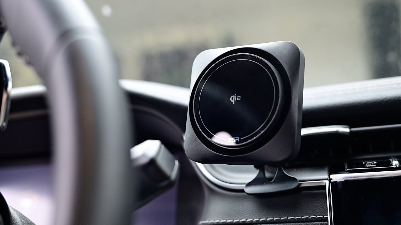 In-car wireless charger mounted on dashboard beside steering wheel, displaying 'qi2' logo, on black surface.