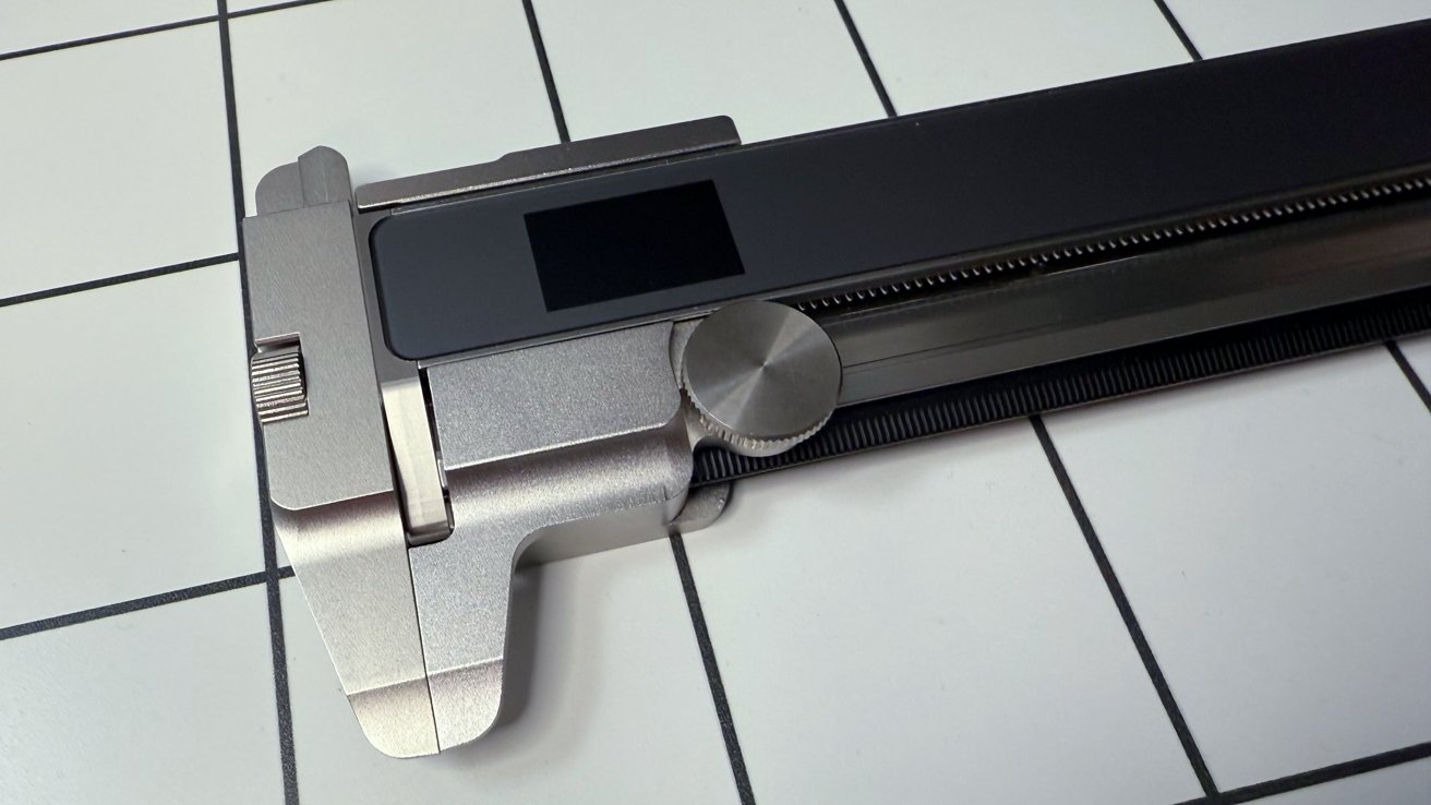 A stainless steel digital caliper with a large dial and screen rests on a tiled surface with thick black lines.