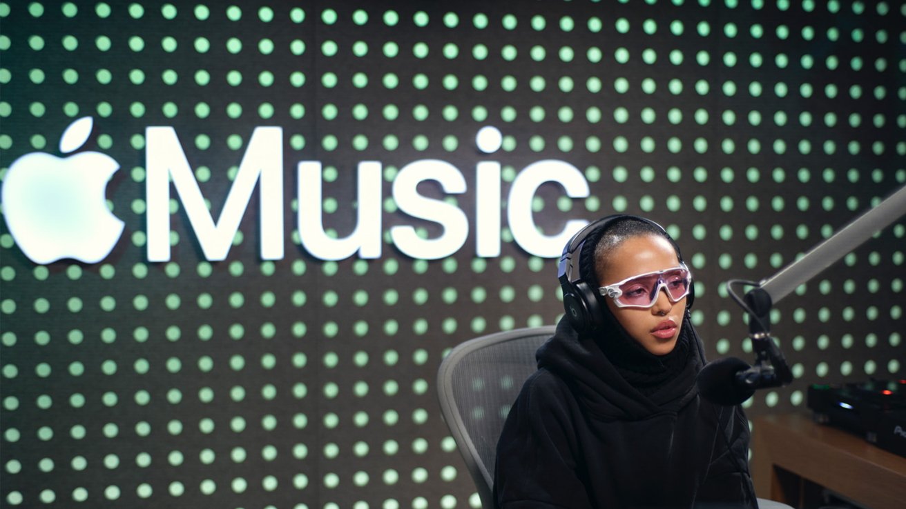 Woman wearing headphones and pink-tinted glasses speaks into a microphone, with an Apple Music logo in the background.