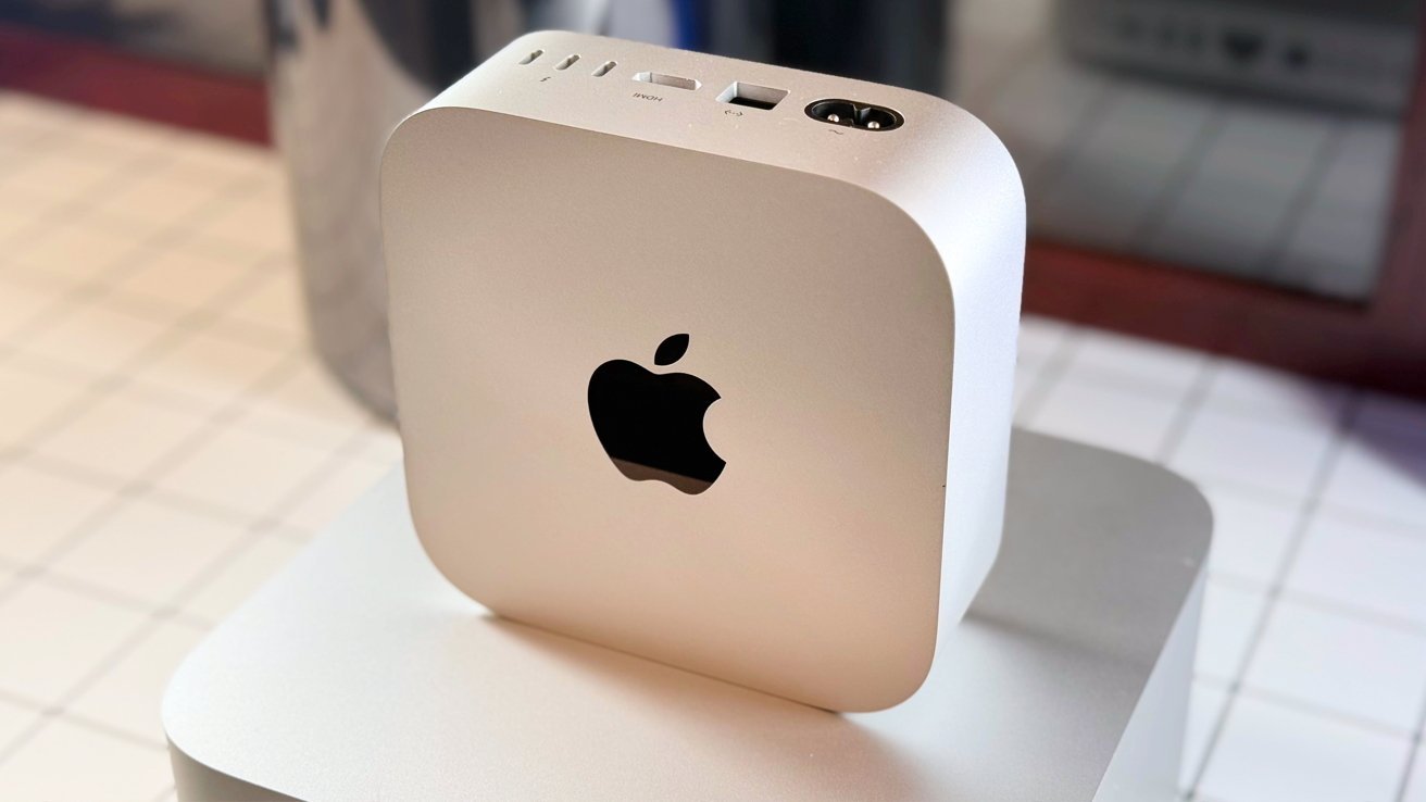 Silver cube-shaped Apple device with logo, rounded corners, and various ports on top, placed on a reflective surface.