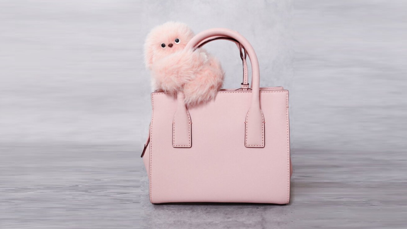 A pink handbag with a cute, fluffy, pink stuffed animal peeking over the handle, set against a gray background.