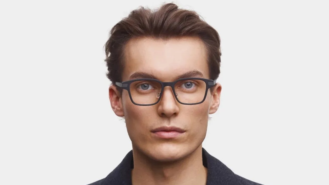 Young man with short brown hair, wearing black rectangular glasses, looking directly ahead, against a plain white background.