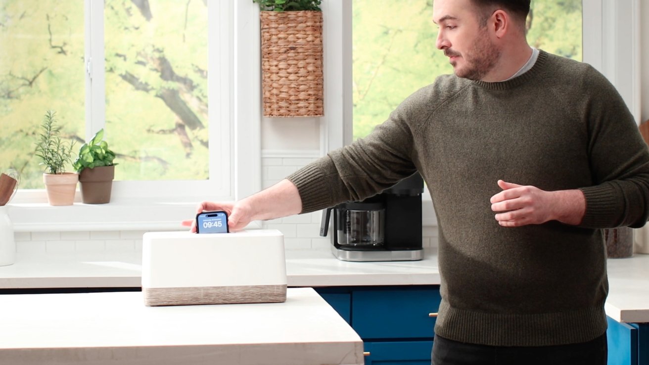 A man in a green sweater places a smartphone displaying 09:45 on a kitchen counter near a coffee maker and potted plants.