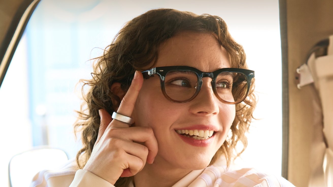 A person with curly hair, wearing glasses and a ring, smiling and sitting inside a vehicle.