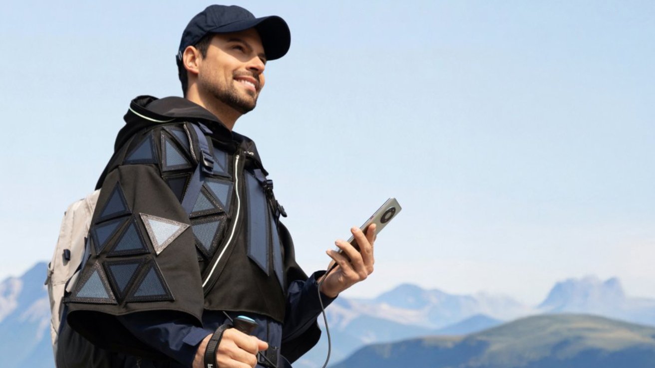 A person wearing triangular-patterned outerwear and a cap holds a smartphone, standing outdoors with mountains in the background.